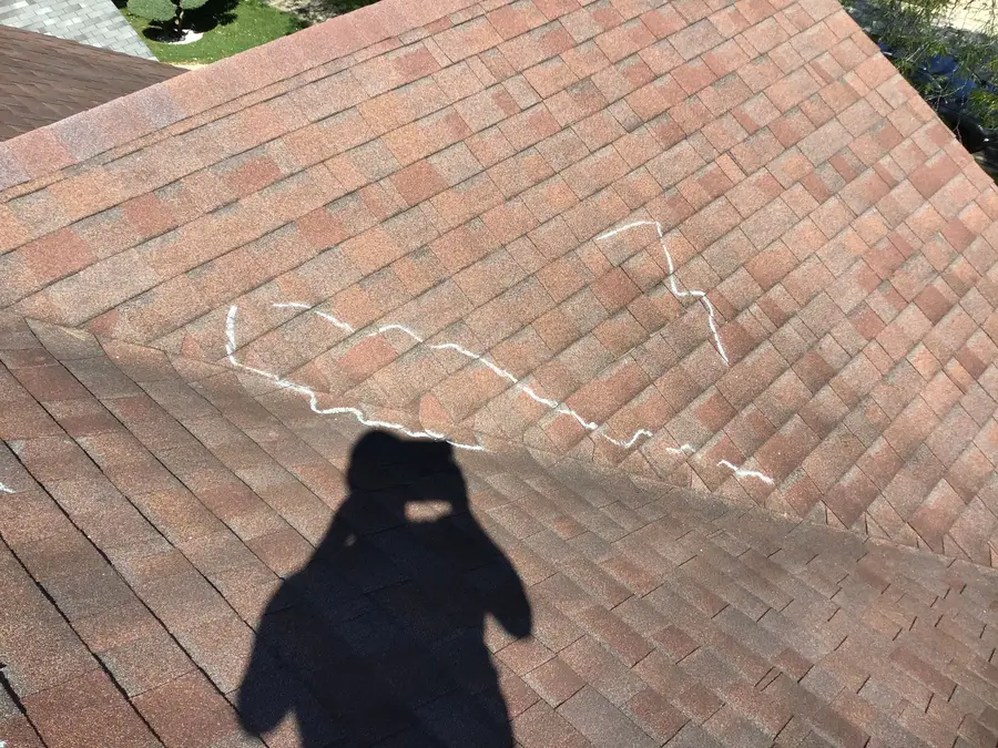 Chalk lines marking hail hits on a reddish brown asphalt shingle slope, with the photographer’s shadow