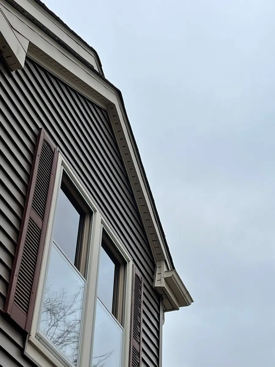 Tall gable wall with tan vinyl siding, a dark shutter and a white window seen from below